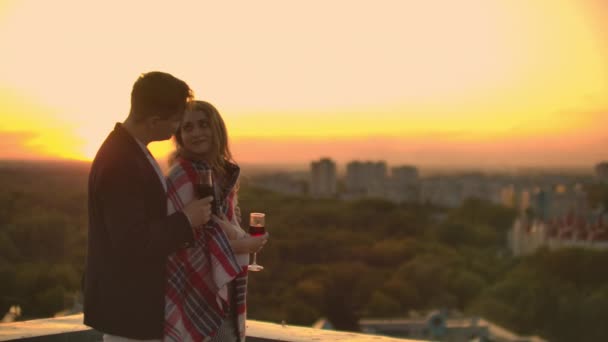 Hombre y mujer bonita joven sentado en el sofá, sosteniendo copas de vino y besándose en la terraza de la azotea al atardecer . — Vídeos de Stock