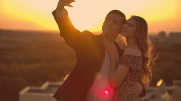 Staande op het dak bij zonsondergang een getrouwd stel een man en een vrouw knuffel en neem een selfie op de telefoon. Maak foto's van jezelf staande op het dak en knuffel. Jonge mensen verliefd. — Stockvideo