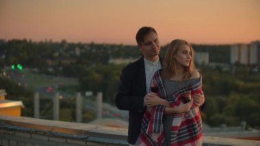 Erkek ve genç güzel kadın kanepede oturan, şarap bardakları tutan ve gün batımında çatı terasında öpüşme.