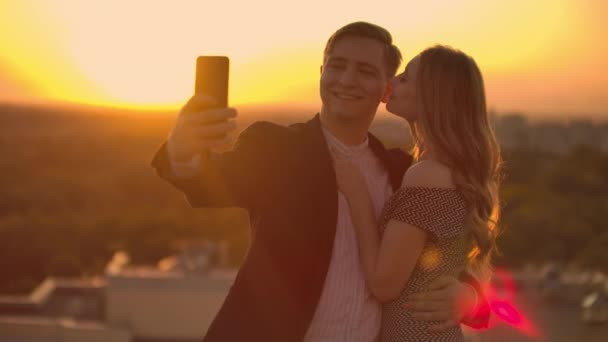 Gün batımında çatıda duran evli bir çift bir erkek ve bir kadın sarılmak ve telefonda selfie çekmek. Çatıda durup kucaklaşırken fotoğraf çek. Aşık gençler. — Stok video