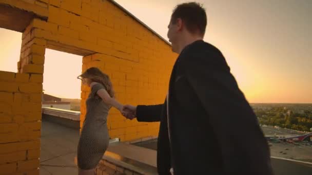 Leuke liefhebbers lachen rennen op het dak van het gebouw bij zonsondergang op de achtergrond van de stad en knuffelen staande aan de rand. Romantiek en liefde. — Stockvideo
