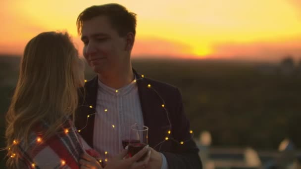 Hombre y mujer bonita joven sentado en el sofá, sosteniendo copas de vino y besándose en la terraza de la azotea al atardecer . — Vídeo de stock