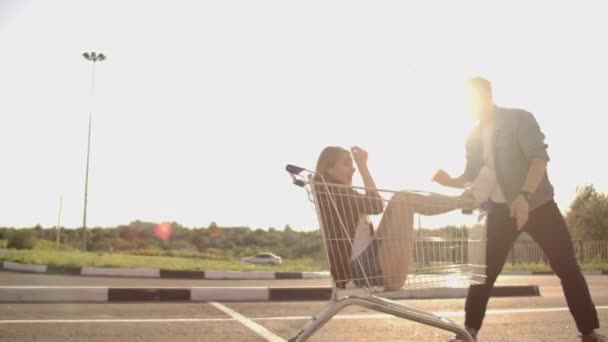 Seitenansicht einer jungen Frau und eines jungen Mannes, die sich im Freien auf Einkaufswagen amüsieren. multiethnische junge Leute, die auf Einkaufswagen rasen. auf dem Parkplatz mit ihren — Stockvideo