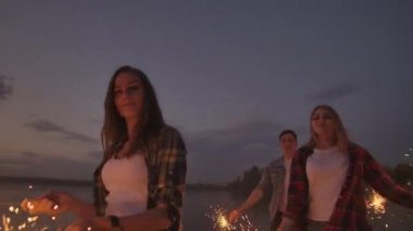 Neşeli erkek ve kadın arkadaşlar gün batımında plaj boyunca koşuyor, yavaş çekimde köpüklü havai fişekler ve kaçak ışıklar tutuyorlar. Sahilde dans ve gün batımı partisi.