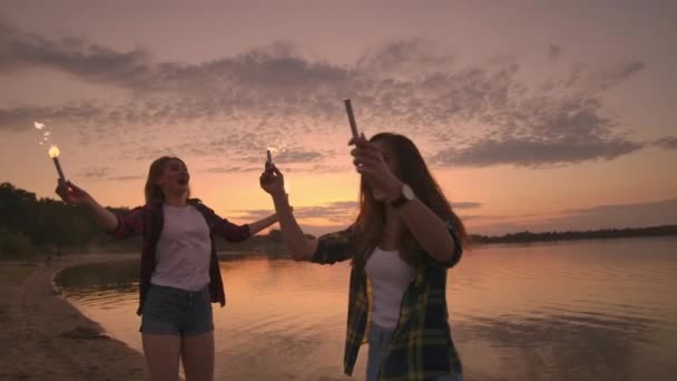 Chicas sonrientes felices corriendo por la playa con bengalas por la noche. Sostén las luces brillantes y ríe. . — Vídeo de stock