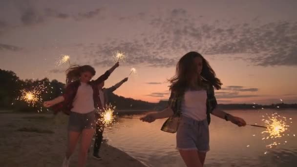 Alegre macho y hembra amigos están corriendo a lo largo de la playa al atardecer, sosteniendo fuegos artificiales brillantes y luces fugitivas en cámara lenta. Fiesta de baile y puesta de sol en la playa . — Vídeo de stock