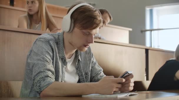 En elev som sitter i ett universitets auditorium med en stor grupp människor sitter i hörlurar och lyssnar på musik och tittar på smartphone-skärmen. Det är i ett tillstånd av lugn. Klassbrytning. — Stockvideo