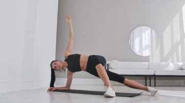 Spor genç seksi esmer kadın beyaz bir iç evde bir eğim ile yan tahta egzersiz yapıyor. Yavaş çekim.