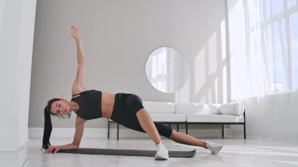 Junge sexy brünette Frau in Sportbekleidung, die zu Hause in einem weißen Interieur seitliche Plankenübung mit einer Neigung macht. Zeitlupe. — Stockvideo