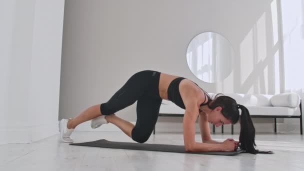 Vrouwelijke brunette fitness trainer demonstreren plank knie naar borst techniek op een mat in huis. — Stockvideo