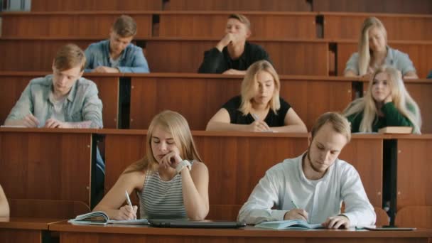 Moderne studenten, mannen en vrouwen, zitten op bureaus in een groot klaslokaal en schrijven een lezing notities. — Stockvideo