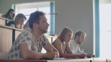 Sıradan kıyafetler giyen kadın ve erkek öğrenciler geniş bir kitlenin içinde bir ders dinler ve ders notları yazarlar.