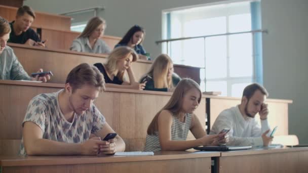 Los estudiantes en una gran audiencia miran las pantallas de los teléfonos inteligentes y no se comunican en la vida real, escriben mensajes solo en línea . — Vídeos de Stock
