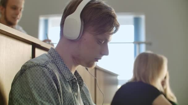 Un estudiante masculino en un auditorio de la Universidad escucha música en auriculares blancos durante un descanso sin prestar atención a los demás. Mucha gente en la audiencia está hablando — Vídeos de Stock