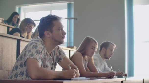 Uomini e donne studenti in abiti ordinari ascoltano una lezione in un vasto pubblico e scrivono note di lezione — Video Stock