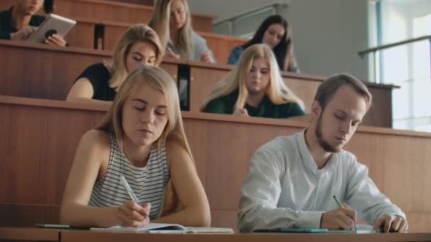 Mężczyźni i kobiety studenci w zwykłych ubrań słuchać wykładu w dużej publiczności i napisać notatki wykładowe — Wideo stockowe