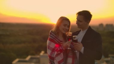 Çatıda genç bir çift bir battaniye içinde duran ve şehir üzerinde güzel gün batımı hayran gözlük şarap içiyor. Şehre bakan çatıda romantik bir akşam.