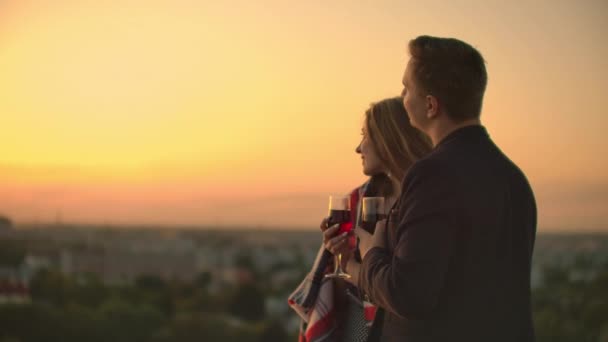 Um jovem casal no telhado bebendo vinho de óculos de pé em um cobertor e admirando o belo pôr do sol sobre a cidade. Noite romântica no telhado com vista para a cidade . — Vídeo de Stock