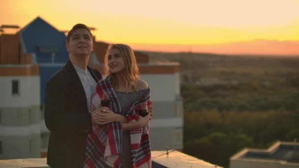 Молода подружня пара на даху обіймає і п'є червоне вино з окулярів, що стоять в одязі в плед і захоплюється прекрасним заходом сонця над містом . — стокове відео
