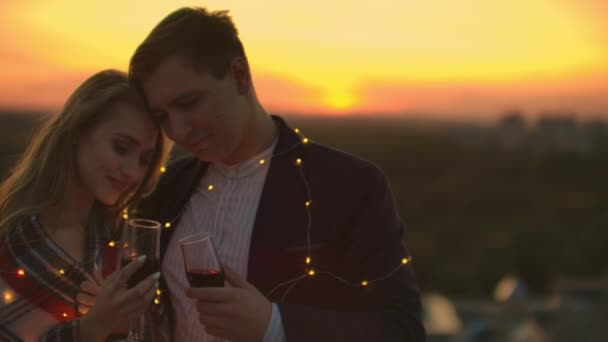 Pareja joven casada en el techo abrazando y bebiendo vino tinto de copas vestidas a cuadros y admirando la hermosa puesta de sol sobre la ciudad . — Vídeos de Stock