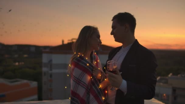 Pareja joven casada en el techo abrazando y bebiendo vino tinto de copas vestidas a cuadros y admirando la hermosa puesta de sol sobre la ciudad . — Vídeos de Stock