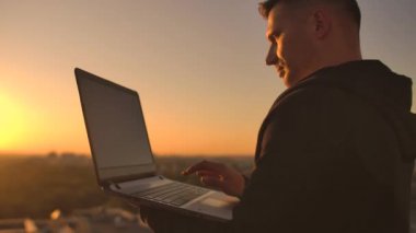 Yakın çekim: Bir programcılar gün batımında çatıya bakan bir dizüstü bilgisayar klavyesi üzerinde el yazma. Bir iş adamı uzaktan çalışır. Freelancer tatilde iş yapar.