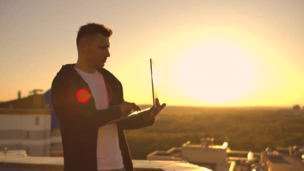 Plan moyen : un pigiste masculin tapant sur un clavier d'ordinateur portable au coucher du soleil surplombant la ville tout en se tenant sur le toit d'un gratte-ciel. L'homme d'affaires travaille à distance et contrôle le travail — Video