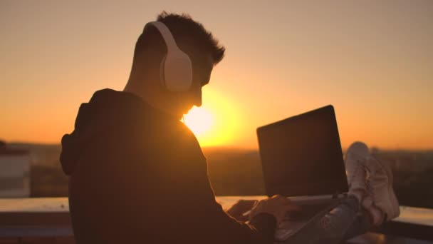 日没時に屋根の上に立つヘッドフォンを持つ男性フリーランサーがキーボードコードページに書き込みます。小さな商売だ音楽を聴き、屋根からの美しい景色を楽しんでコンピュータで仕事 — ストック動画