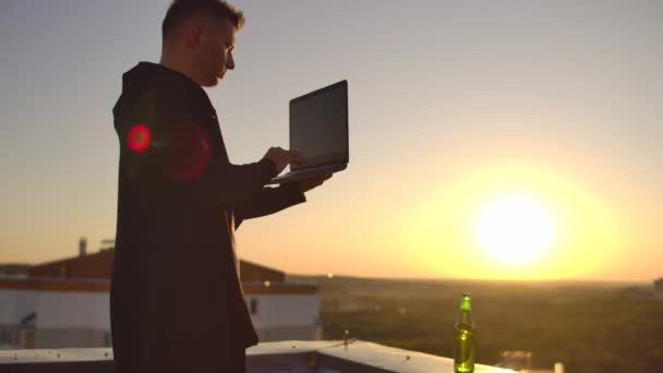 Un corredor de bolsa freelancer masculino se para en una azotea al atardecer con una computadora portátil y escribe en un teclado con los dedos mirando el paisaje urbano desde una vista de pájaro — Vídeos de Stock