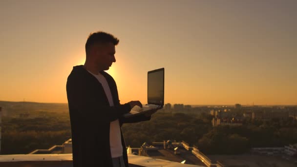 Um corretor de bolsa freelancer masculino fica em um telhado ao pôr do sol com um laptop e digita em um teclado com os dedos olhando para a paisagem urbana de uma visão de olhos de pássaros — Vídeo de Stock