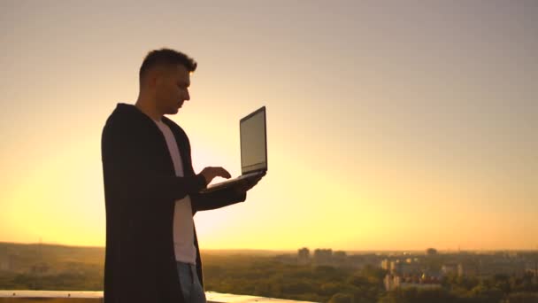 Un corredor de bolsa freelancer masculino se para en una azotea al atardecer con una computadora portátil y escribe en un teclado con los dedos mirando el paisaje urbano desde una vista de pájaro — Vídeo de stock