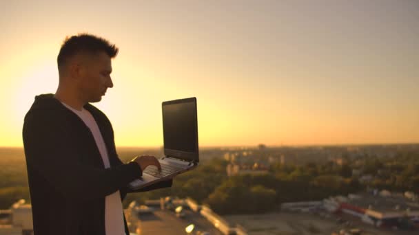 Un programador freelance masculino se sienta en un techo de rascacielos con una computadora portátil y un código de escritura de cerveza en un teclado durante la puesta del sol. Trabajo remoto . — Vídeo de stock