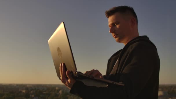 Programador um hacker está no telhado com um laptop ao pôr do sol diz código de erro no teclado e olhando para a vista da cidade — Vídeo de Stock