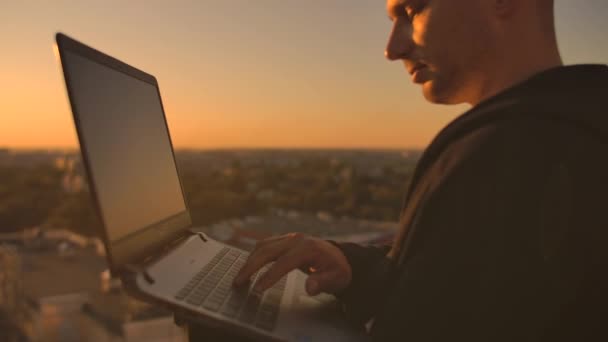 Un corredor de bolsa freelancer masculino se para en una azotea al atardecer con una computadora portátil y escribe en un teclado con los dedos mirando el paisaje urbano desde una vista de pájaro — Vídeo de stock