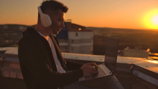 Ein Mann mit Kopfhörern, der bei Sonnenuntergang auf dem Dach steht, schreibt auf die Tastatur-Codeseiten. wenig Geschäft. Musik hören und am Computer arbeiten und die schöne Aussicht vom Dach genießen — Stockvideo