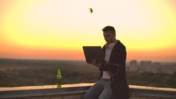 Человек в наушниках на крыше расслабляется, работая удаленно наслаждаясь жизнью, несмотря на красивый вид потягивая пиво и типы на клавиатуре . — стоковое видео