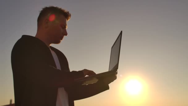 Plano medio: un freelancer masculino escribiendo en un teclado portátil al atardecer con vistas a la ciudad mientras está de pie en el techo de un rascacielos. El empresario trabaja a distancia y controla el trabajo — Vídeos de Stock