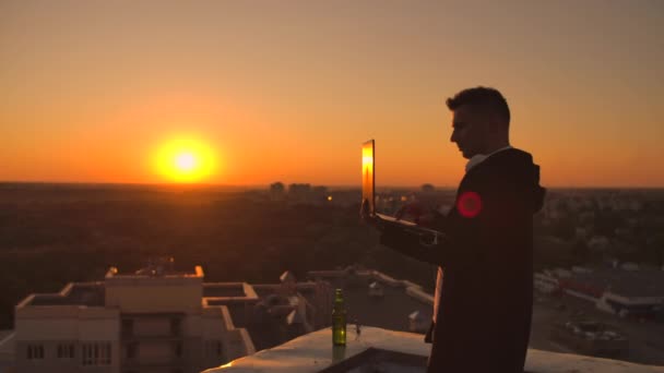 Un corredor de bolsa freelancer masculino se para en una azotea al atardecer con una computadora portátil y escribe en un teclado con los dedos mirando el paisaje urbano desde una vista de pájaro — Vídeos de Stock