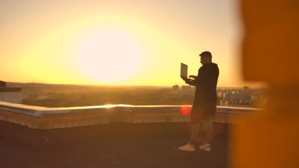 Um corretor de bolsa freelancer masculino fica em um telhado ao pôr do sol com um laptop e digita em um teclado com os dedos olhando para a paisagem urbana de uma visão de olhos de pássaros — Vídeo de Stock