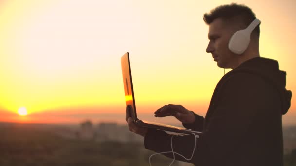Een man freelancer in hoofdtelefoons staande op het dak bij zonsondergang schrijft op de toetsenbord codepagina's. Weinig zaken. Luister naar muziek en werk op de computer genietend van het prachtige uitzicht vanaf het dak — Stockvideo