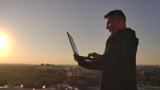Programista haker jest na dachu z laptopem o zachodzie słońca mówi kod błędu na klawiaturze i patrząc na widok na miasto — Wideo stockowe