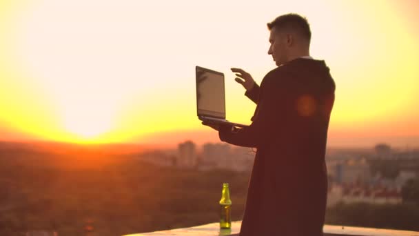 Un corredor de bolsa freelancer masculino se para en una azotea al atardecer con una computadora portátil y escribe en un teclado con los dedos mirando el paisaje urbano desde una vista de pájaro — Vídeos de Stock