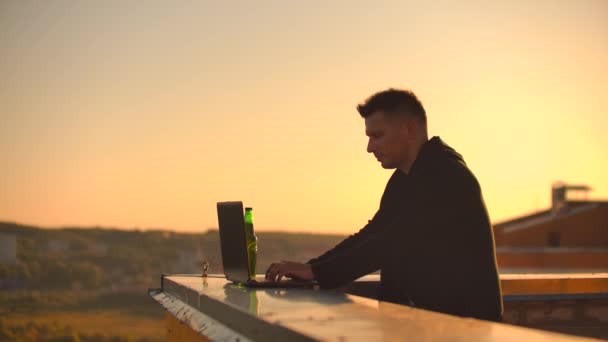 Чоловічий позаштатний програміст сидить на даху хмарочоса з ноутбуком і кодом для друку пива на клавіатурі під час заходу сонця. Віддалена робота . — стокове відео