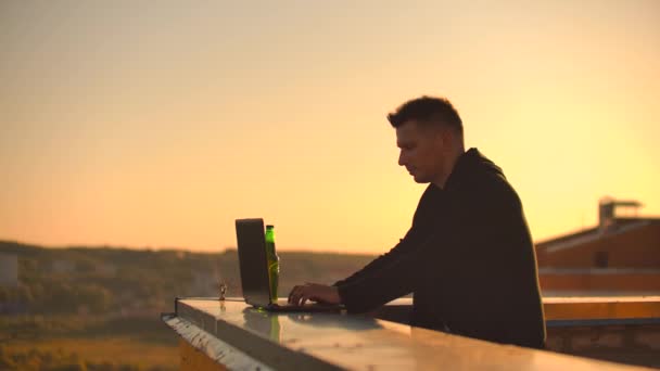 Un programador freelance masculino se sienta en un techo de rascacielos con una computadora portátil y un código de escritura de cerveza en un teclado durante la puesta del sol. Trabajo remoto . — Vídeo de stock