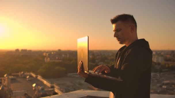 中间计划：一名男性自由职业者在日落时分在笔记本电脑键盘上打字，站在摩天大楼的屋顶上，俯瞰城市。商人远程工作并控制工作 — 图库视频影像