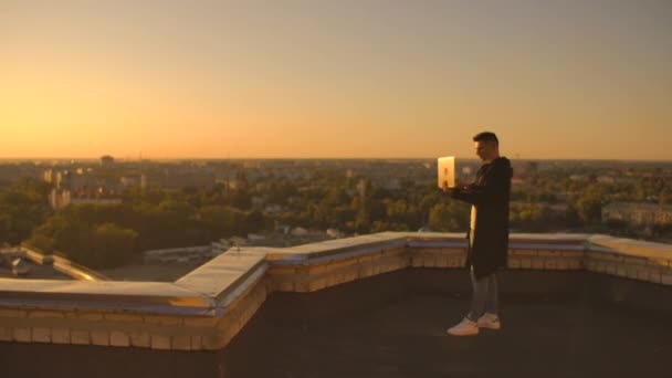 Piano intermedio: un freelance di sesso maschile che digita su una tastiera portatile al tramonto con vista sulla città mentre si trova sul tetto di un grattacielo. L'uomo d'affari lavora a distanza e controlla il lavoro — Video Stock