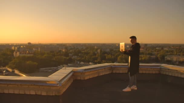 Middle plan: een mannelijke freelancer typen op een laptop toetsenbord bij zonsondergang met uitzicht op de stad terwijl staan op het dak van een wolkenkrabber. De zakenman werkt op afstand en regelt het werk — Stockvideo