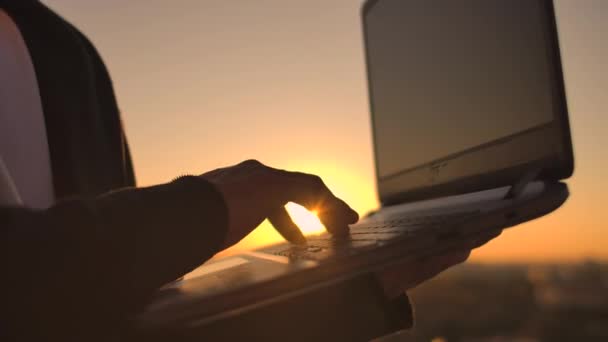 Primo piano: un programmatore digita a mano su una tastiera portatile al tramonto con vista sul tetto. Un uomo d'affari lavora a distanza. Freelancer svolge il lavoro in vacanza . — Video Stock