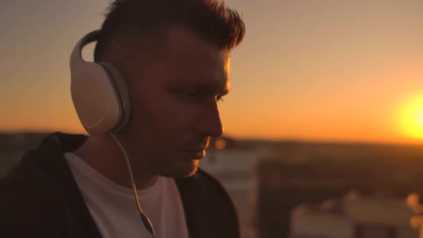 Um homem freelancer em fones de ouvido em pé no telhado ao pôr do sol escreve nas páginas de código do teclado. Pequenos negócios. Ouça música e trabalhe no computador apreciando a bela vista do telhado — Vídeo de Stock