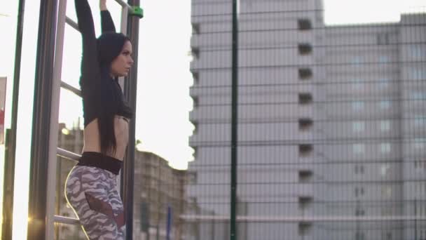 Slow motion: Candid shot of real healthy and fit woman performing hanging leg raises on outdoor fitness station in sunset at city park. Mostrando un fuerte paquete de seis abdominales . — Vídeos de Stock